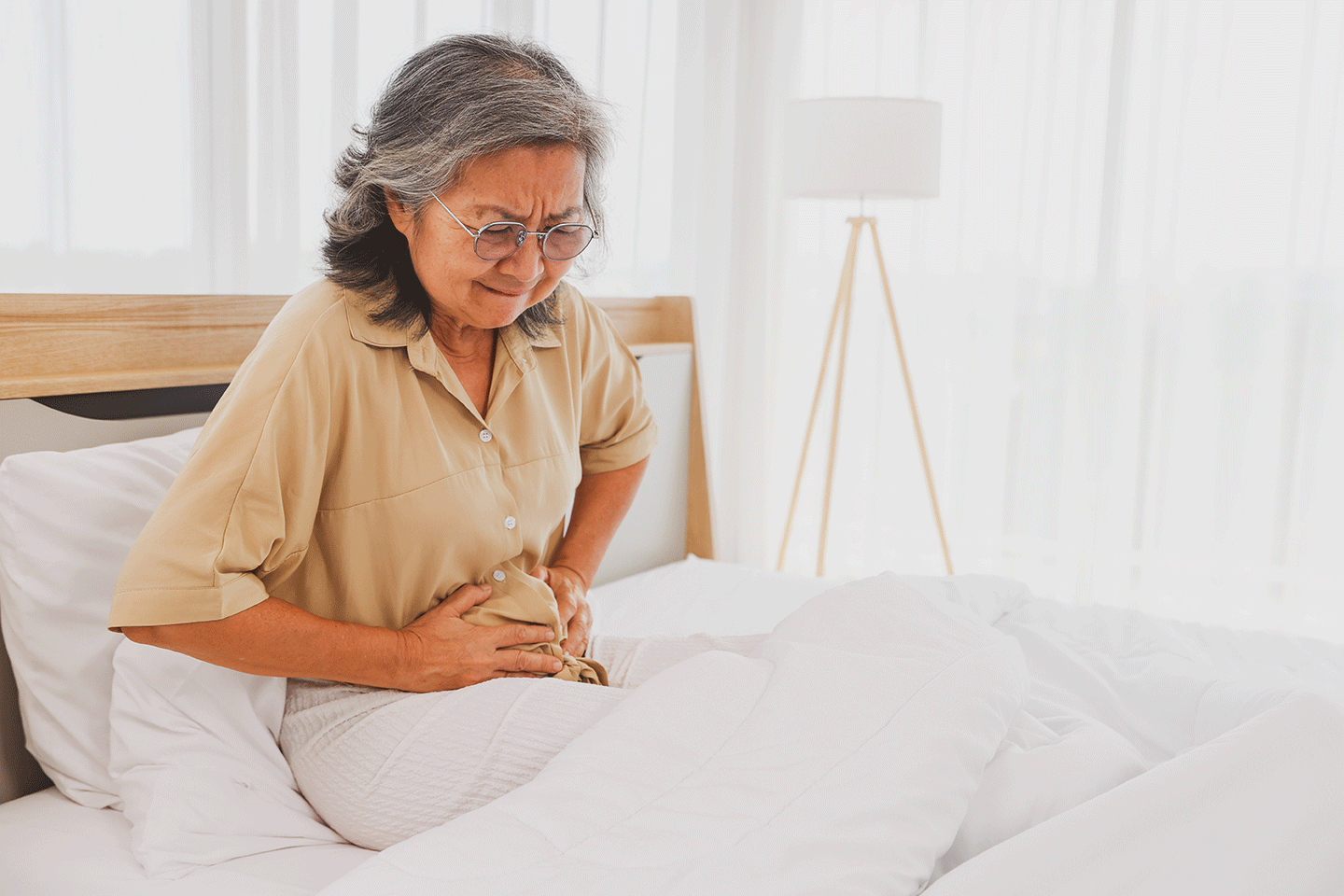 Elderly Asian woman sitting and holding her abdomen with a distressed expression, illustrating stomach pain and digestive discomfort associated with colon conditions.